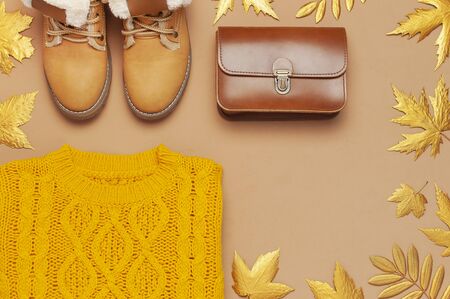 Brown leather women bag, orange knitted sweater, warm boots, golden autumn leaf on brown background top view flat lay. Fashionable women's accessories. Autumn Fashion Concept. Stylish Lady Clothes.の写真素材