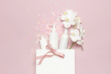 Flat lay White cosmetic bottle containers festive confetti gift bag White Phalaenopsis orchid flowers on pink background top view. Cosmetics SPA branding mock-up Natural organic beauty product.の写真素材