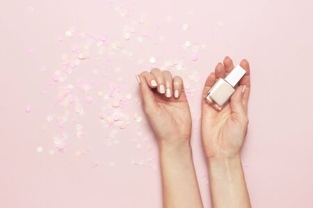Stylish fashionable female manicure in pastel colors. Hands of young girl holding bottle of nail polish on pink background with festive confetti. Minimalist manicure trend Flat lay top view copy space.の写真素材