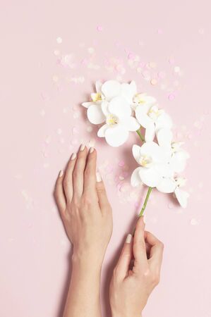 Woman's hands with gentle manicure hold branch of White Phalaenopsis orchid flowers on pastel pink background with festive confetti top view flat lay. Tropical flower. Holiday Women's Day Flower Card.の写真素材