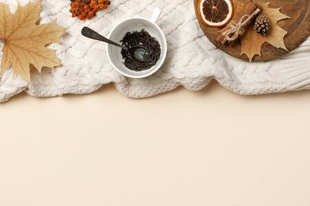 Flat lay autumn composition. White cup with tea leaves, wooden plate, knitted fashion sweater or plaid, yellow maple leaves, cinnamon, dry mountain ash, orange on beige background top view copy space.の写真素材