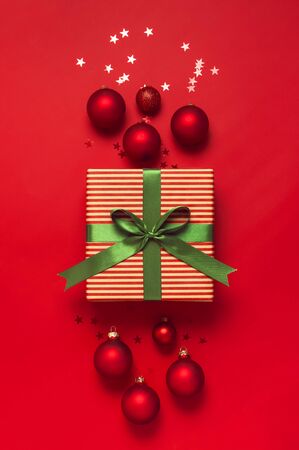 Christmas background. Gift with green ribbon, red Christmas balls, confetti shape of stars on red background top view. New Year presents Festive decorations Flat lay Xmas holiday 2020 celebration.の写真素材