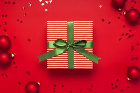Christmas background. Gift with green ribbon, red Christmas balls, confetti shape of stars on red background top view. New Year presents Festive decorations Flat lay Xmas holiday 2020 celebration.の写真素材