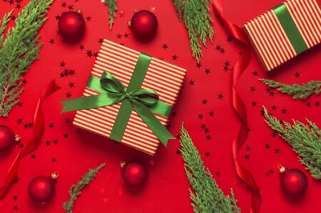 Christmas background. Gift with green ribbon, red balls, christmas tree branches, confetti stars on red background top view. New Year presents Festive decorations Flat lay Xmas holiday 2020 celebration.の写真素材