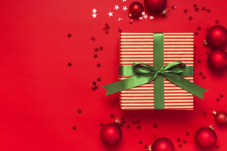 Christmas background. Gift with green ribbon, red Christmas balls, confetti shape of stars on red background top view. New Year presents Festive decorations Flat lay Xmas holiday 2020 celebration.の写真素材