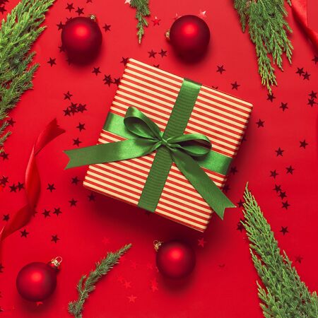 Christmas background. Gift with green ribbon, red balls, christmas tree branches, confetti stars on red background top view. New Year presents Festive decorations Flat lay Xmas holiday 2020 celebration.の写真素材