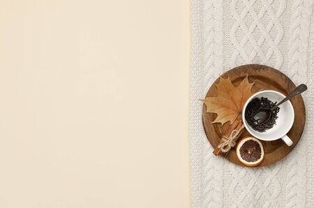 Flat lay autumn composition. White cup with tea leaves, wooden plate, knitted fashion sweater or plaid, yellow maple leaves, cinnamon, dry mountain ash, orange on beige background top view copy space.の写真素材
