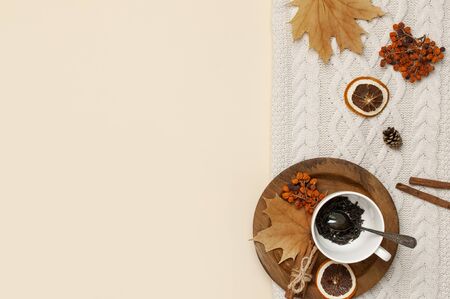 Flat lay autumn composition. White cup with tea leaves, wooden plate, knitted fashion sweater or plaid, yellow maple leaves, cinnamon, dry mountain ash, orange on beige background top view copy space.の写真素材