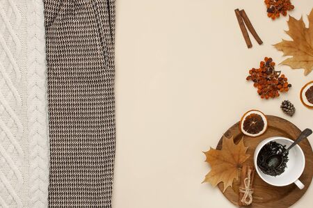 Flat lay autumn composition. White cup with tea leaves, wooden plate, knitted fashion sweater or plaid, pants in a pattern, yellow maple leaves, cinnamon, orange on beige background top view copy space.の写真素材
