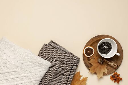 Flat lay autumn composition. White cup with tea leaves, wooden plate, knitted fashion sweater or plaid, pants in a pattern, yellow maple leaves, cinnamon, orange on beige background top view copy space.の写真素材