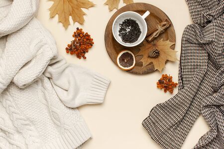 Flat lay autumn composition. White cup with tea leaves, wooden plate, knitted fashion sweater or plaid, pants in a pattern, yellow maple leaves, cinnamon, orange on beige background top view copy space.の写真素材