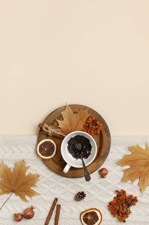 Flat lay autumn composition. White cup with tea leaves, wooden plate, knitted fashion sweater or plaid, yellow maple leaves, cinnamon, dry mountain ash, orange on beige background top view copy space.の写真素材