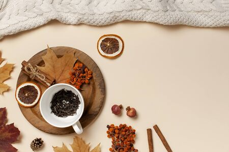 Flat lay autumn composition. White cup with tea leaves, wooden plate, knitted fashion sweater or plaid, yellow maple leaves, cinnamon, dry mountain ash, orange on beige background top view copy space.の写真素材