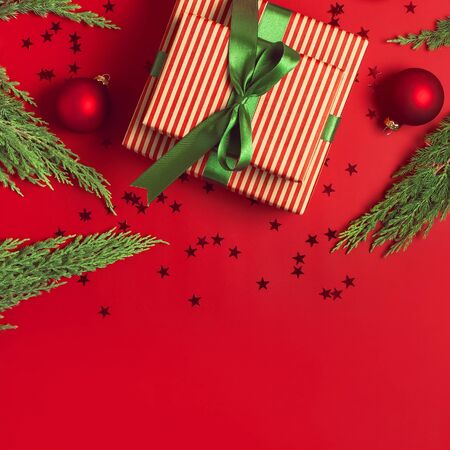 Christmas background. Gift with green ribbon, red balls, christmas tree branches, confetti stars on red background top view. New Year presents Festive decorations Flat lay Xmas holiday 2020 celebration.の写真素材
