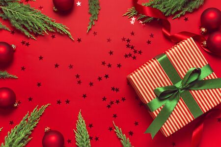 Christmas background. Gift with green ribbon, red balls, christmas tree branches, confetti stars on red background top view. New Year presents Festive decorations Flat lay Xmas holiday 2020 celebration.の写真素材