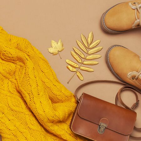 Brown leather women bag, orange knitted sweater, warm boots, golden autumn leaf on brown background top view flat lay. Fashionable womens accessories. Autumn Fashion Concept. Stylish Lady Clothes.の写真素材