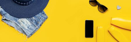 Female summer fashion background. Clothes and accessories on yellow background. Blue hat, denim shorts, sandals, sunglasses, sunscreen, phone. Flat lay top view, beauty or fashion blog concept.の写真素材
