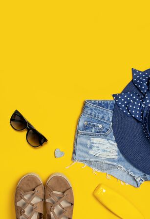 Female summer fashion background. Clothes and accessories on yellow background. Blue hat, denim shorts, sandals, sunglasses, sunscreen, phone. Flat lay top view, beauty or fashion blog concept.の写真素材