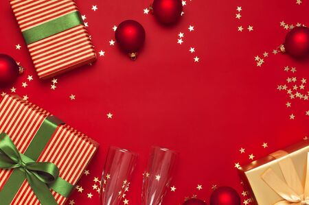 New Year Christmas presents, Christmas balls, champagne glasses, gold confetti stars on red background top view. Flat lay Xmas holiday 2020 celebration. Gift boxes greeting card Festive decorations.の写真素材
