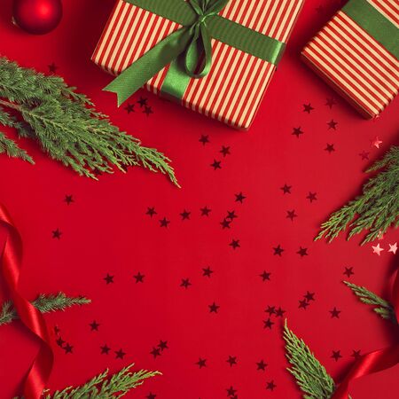 Christmas background. Gift with green ribbon red balls christmas tree branches confetti stars on red background top view. New Year presents Festive decorations Flat lay Xmas holiday 2020 celebration.の写真素材