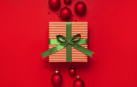 Christmas background. Gift with green ribbon, red Christmas balls, confetti shape of stars on red background top view. New Year presents Festive decorations Flat lay Xmas holiday 2020 celebration.の写真素材