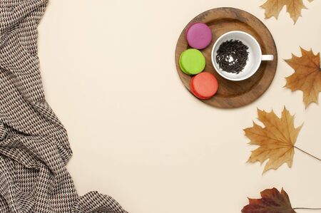 Flat lay autumn composition. White cup with tea leaves, wooden plate, colorful cakes macaroon, pants in a pattern, yellow maple leaves cinnamon orange on beige background top view copy space.の写真素材