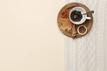 Flat lay autumn composition. White cup with tea leaves, wooden plate, knitted fashion sweater or plaid, yellow maple leaves, cinnamon, dry mountain ash, orange on beige background top view copy space.の写真素材