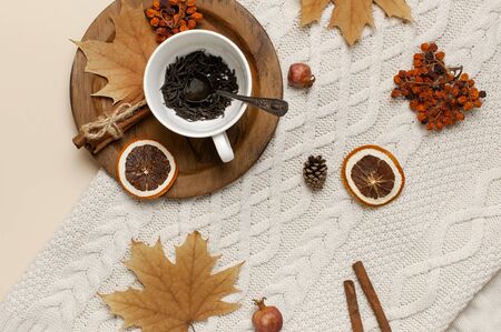 Flat lay autumn composition. White cup with tea leaves, wooden plate, knitted fashion sweater or plaid, yellow maple leaves, cinnamon, dry mountain ash, orange on beige background top view copy space.の写真素材