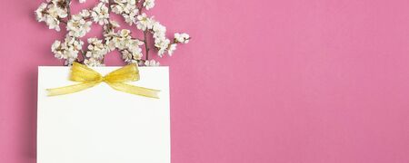 White gift bag with gold ribbon and branch of spring white flowers on bright pink background. Greeting card with delicate flowers Pink floral background. Spring minimal concept. Flat lay top view.の写真素材