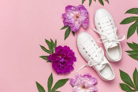 White female fashion sneakers and pink purple flowers peonies on pink background. Flat lay top view copy space. Women's shoes. Stylish white sneakers. Fashion blog or magazine. Spring shoe concept.の写真素材