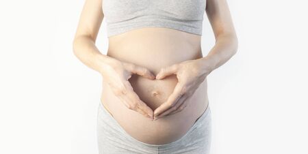 Beautiful young pregnant woman holding hands symbol heart on her belly on white background. The concept of expectation of the child, pregnancy and motherhood, happy baby waiting. Tender photo.の写真素材