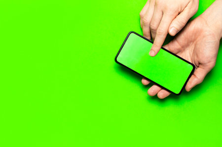 Male hands hold a modern black smartphone with green blank screen on neon green background flat lay...