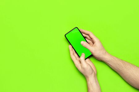Male hands hold a modern black smartphone with green blank screen on neon green background flat lay top view. Modern technology, phone, gadget in hands, touch screen, template for your design. Mockup.の写真素材