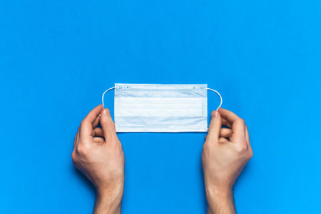 Medical mask in male hands on blue background flat lay top view. Protection against virus...