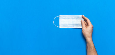 Medical mask in male hands on blue background flat lay top view. Protection against virus...