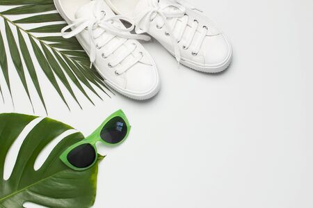 White female fashion sneakers, trendy sunglasses, tropical leaves on light gray background. Flat lay top view copy space. Women's shoes, accessories. Stylish white sneakers. Creative summer concept.の写真素材