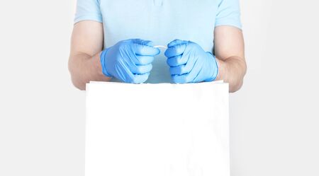 Concept of delivery of goods during quarantine. Young man in blue gloves holds white paper bag in his hands on white background. Delivery against Coronavirus 2019-nCov in pandemic Contactless delivery.の写真素材