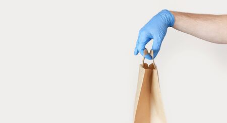 Male hand in blue medical gloves holds craft paper bag on white background. Delivery against Coronavirus 2019-nCov in pandemic Contactless delivery. Concept of delivery of goods during quarantine.の写真素材