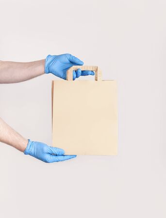 Delivery against Coronavirus 2019-nCov in pandemic Contactless delivery. Male hand in blue medical gloves holds craft paper bag on white background. Concept of delivery of goods food during quarantine.の写真素材