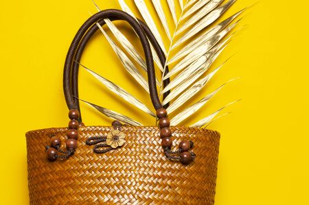 Beach accessories summer bag. Beach wicker straw or rattan women's eco bag with golden tropical palm leaf on yellow background. Flat lay top view copy space. Concept of travel Summer bright background.の写真素材