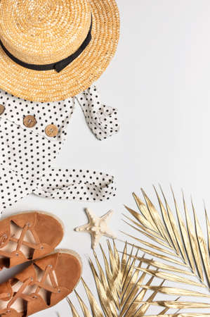 Summer womens white dress in black peas brown sandals straw hat golden palm leaf shells starfish on light background. Flat lay top view copy space. Womens beach fashion travel vacation golden shades.の写真素材