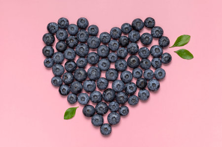 Fresh juicy blueberries with green leaves in the shape of heart on pink background. Blueberries...