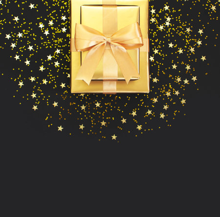 Golden gift with ribbon, confetti stars on black background. Flat lay, top view, copy space. Golden decorations, party, birthday, holiday concept. Gift box, surprise. New Year or Christmas composition.の写真素材