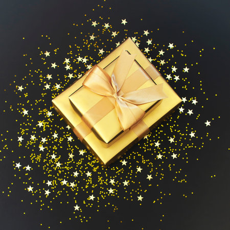 Golden gift with ribbon, confetti stars on black background. Flat lay, top view, copy space. Golden...
