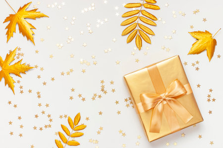 Flat lay Autumn holiday concept. Golden gift or present box with ribbon, confetti stars, golden autumn leaves on light gray background top view copy space. Surprise congratulations birthday.の写真素材