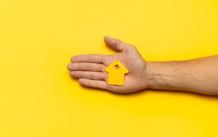 House symbol in male hands on yellow background flat lay top view. Model of plastic yellow house....
