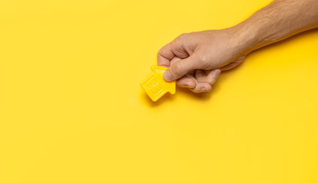 House symbol in male hands on yellow background flat lay top view. Model of plastic yellow house....