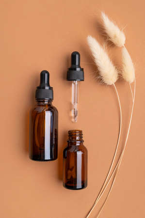 Clean brown glass cosmetic bottles with pipette, jar of cream, decorative spikelets plants on brown background. natural organic cosmetics. Spa products for health and beauty. Serum, facial oil.の写真素材