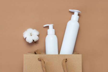 Cosmetics SPA branding mock-up. White cosmetic bottle containers, cotton flowers, paper craft bag on brown background top view flat lay. Natural organic beauty product concept.の写真素材