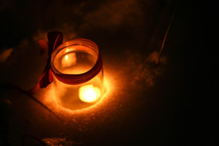 A small candle burns in a jar in the dark against the background of bokeh lights red ribbon bow, bokeh, blurred background, place for textの写真素材